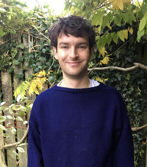 Photo of Alex Laar smiling to camera. He is standing outside, in front of an ivy covered fence. Foliage and a tree branch is visible behind.