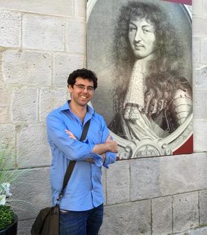 Photo of Jonathan Healey smiling, leaning against a wall with a large reproduction of an early modern engraved portrait. 