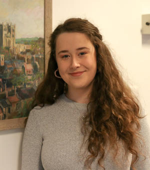 Photo of Clare Burgess smiling to camera, standing in front of a painting of a city. 