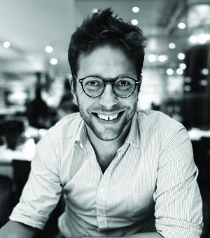 Black and white photo of Olly Markeson. Wearing white shirt and glasses. Blurred restaurant background.